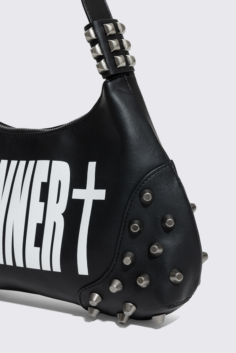 Black faux leather shoulder bag with white sinner cross print, studded panels and grunge aesthetic.