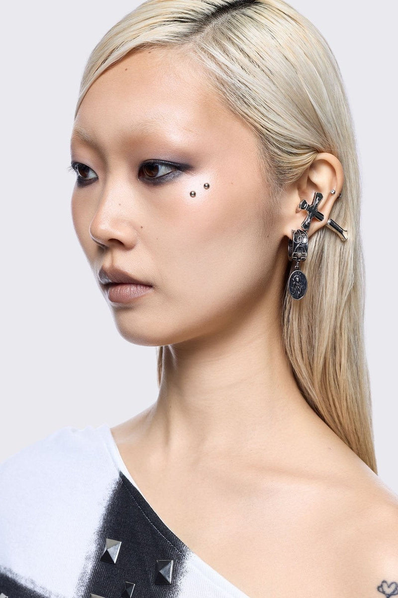 A 5-piece set of mismatched silver-toned earrings and an ear cuff featuring gothic and religious motifs, including crosses, a Virgin Mary medallion, and a winged sword dangle.