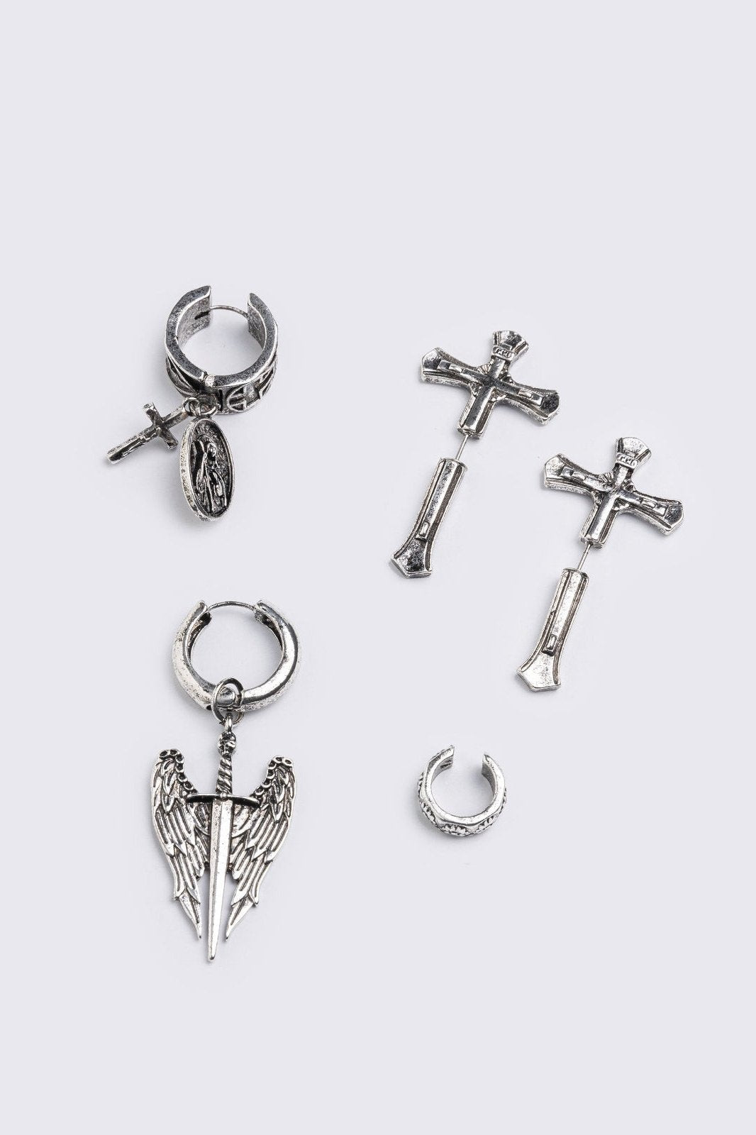 A 5-piece set of mismatched silver-toned earrings and an ear cuff featuring gothic and religious motifs, including crosses, a Virgin Mary medallion, and a winged sword dangle.