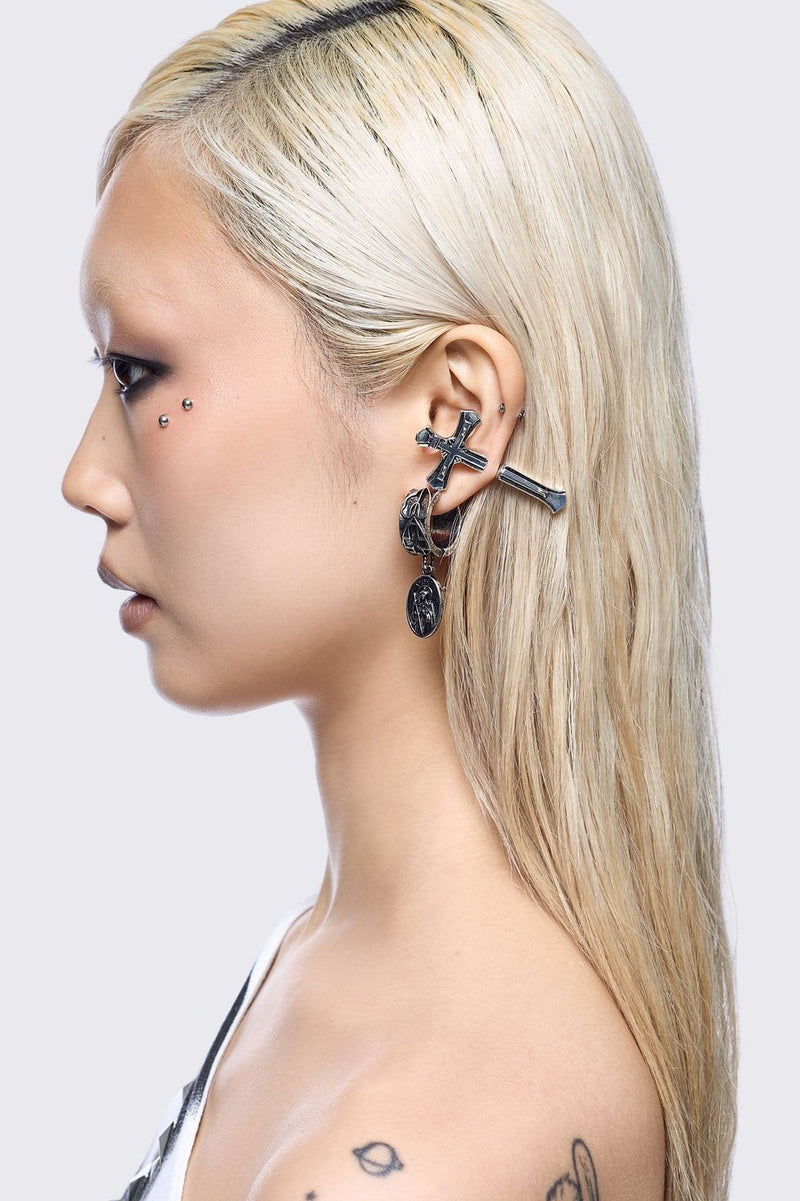 A 5-piece set of mismatched silver-toned earrings and an ear cuff featuring gothic and religious motifs, including crosses, a Virgin Mary medallion, and a winged sword dangle.