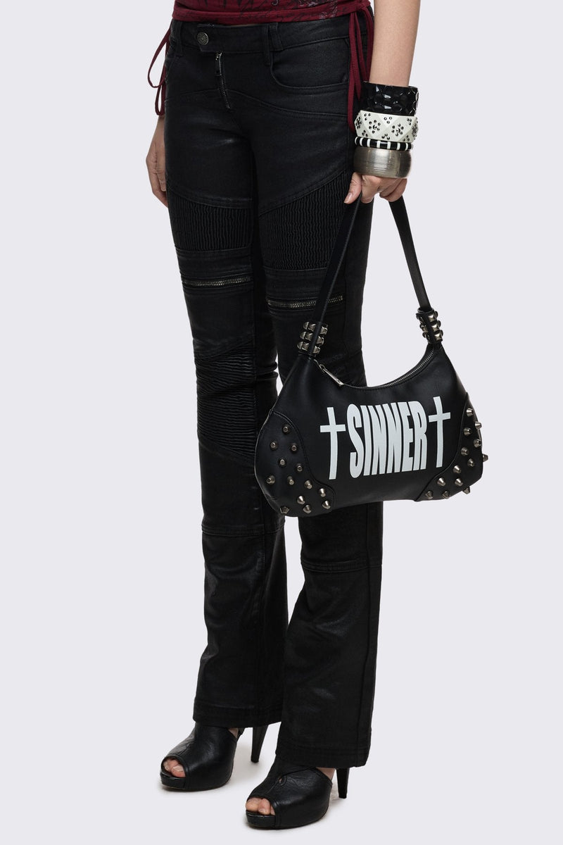 Black faux leather shoulder bag with white sinner cross print, studded panels and grunge aesthetic.